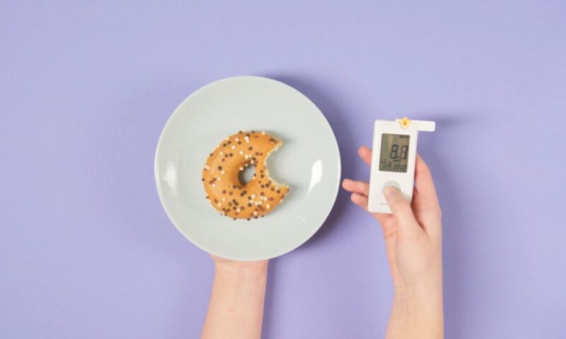 prikaz krofne i aparata za merenje šećera na ljubičastoj pozadini